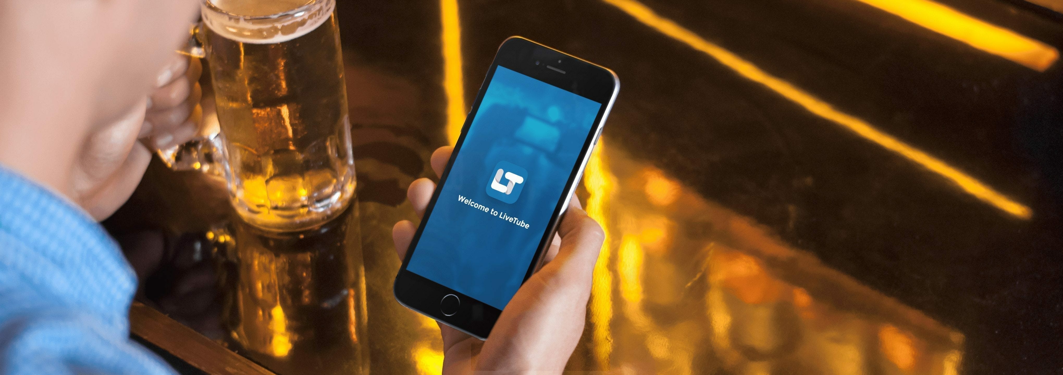LiveTube - A person sits at a reflective table, a beer mug nearby, holding a smartphone displaying both a rideshare app interface and options on how to make money live streaming.