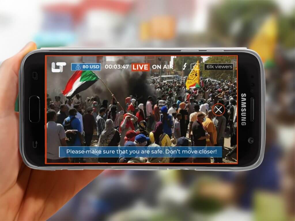 一隻手拿著智慧手機,顯示 LIVETUBE 應用程式,顯示以旗幟和煙霧為背景的擁擠抗議現場的現場直播。一條安全資訊寫道:“請確保您的安全。不要靠近! 一隻手拿著智慧手機,顯示 LIVETUBE 應用程式,顯示以旗幟和煙霧為背景的擁擠抗議現場的現場直播。一條安全資訊寫道:“請確保您的安全。不要靠近!