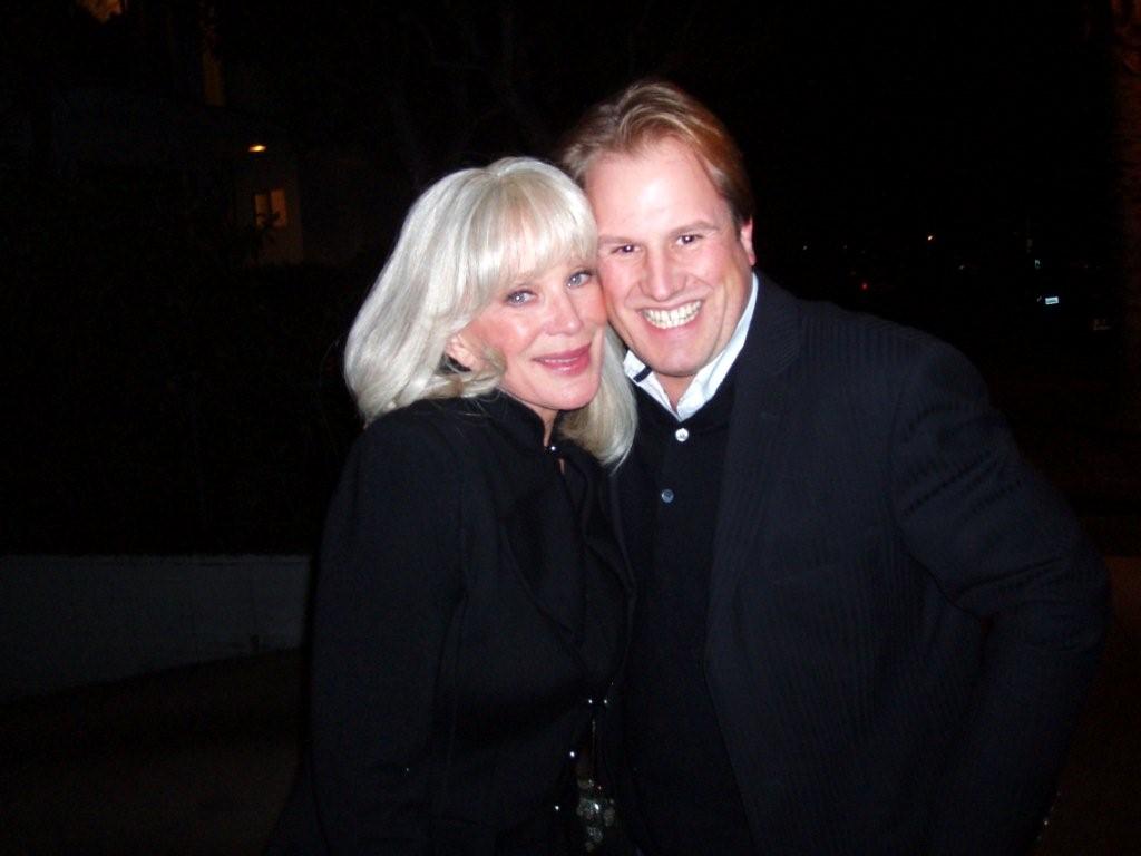 LiveTube - Two people, a woman with long white hair and a man with light brown hair, stand close together and smile at the camera at night.