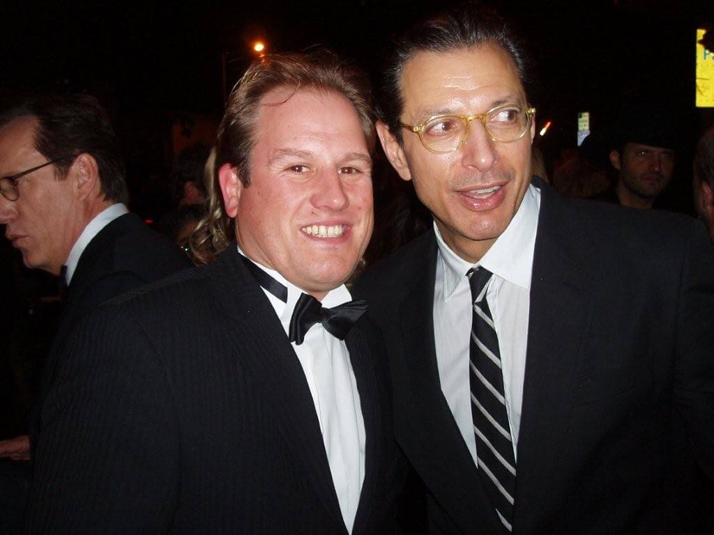 LiveTube - Two men in suits and ties pose together and smile at an event; other people are visible in the background.