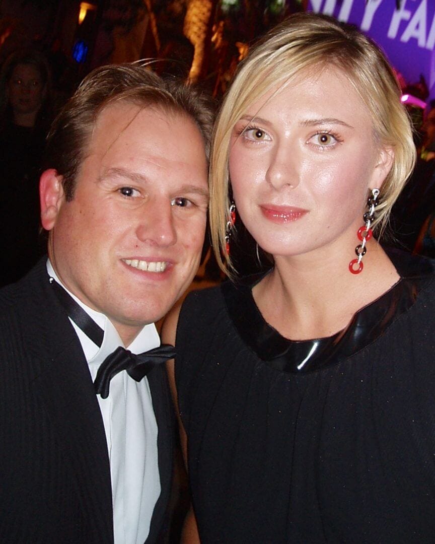LiveTube - A man in a tuxedo and a woman in a black dress pose together at an indoor event with colorful lighting in the background.