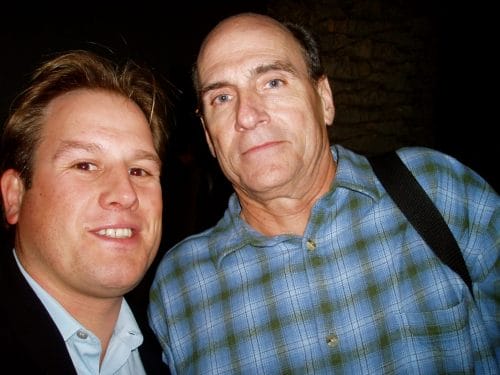 LiveTube - Two men pose for a close-up photo together; one is wearing a blue shirt and the other is in a suit jacket and collared shirt.