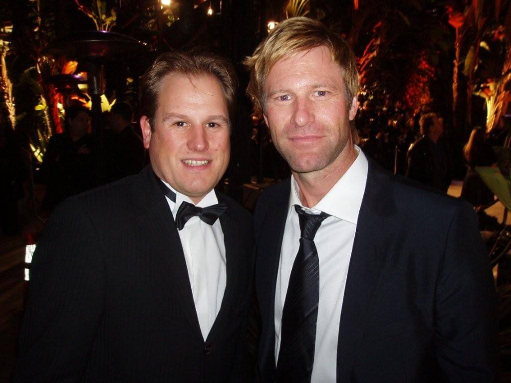 LiveTube - Two men in suits, one wearing a bow tie and the other a tie, pose together at an evening event with blurred people and lights in the background.