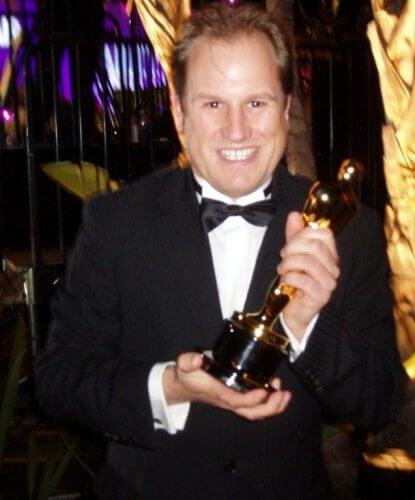LiveTube - A man in a black tuxedo smiles while holding an Oscar trophy in both hands at an indoor event.