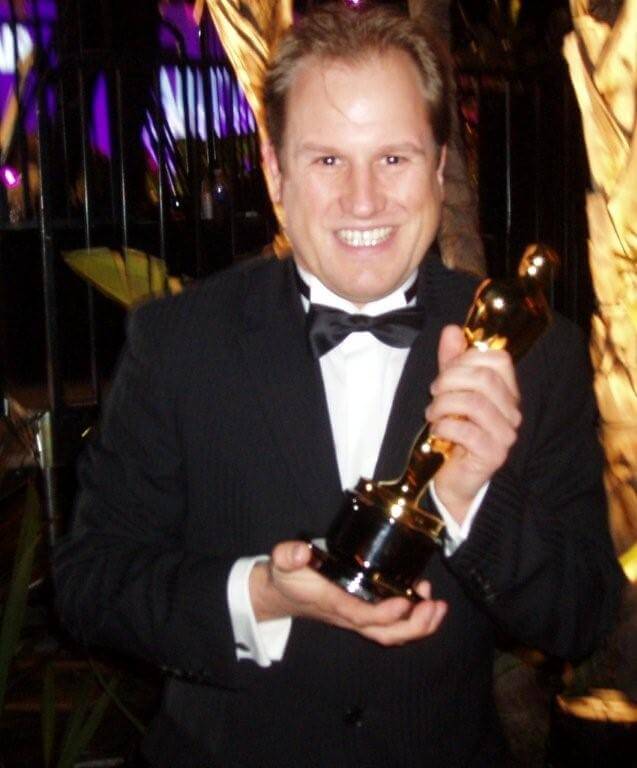 LiveTube - A man in a black tuxedo smiles while holding an Oscar trophy in both hands at an indoor event.