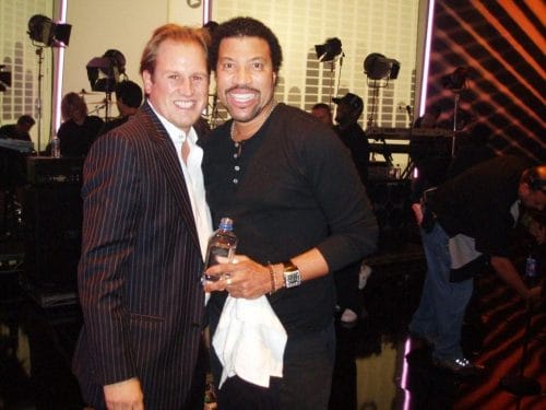 LiveTube - Two men stand together smiling at the camera in a studio setting; one holds a water bottle and a white towel. Studio lights and crew are visible in the background.