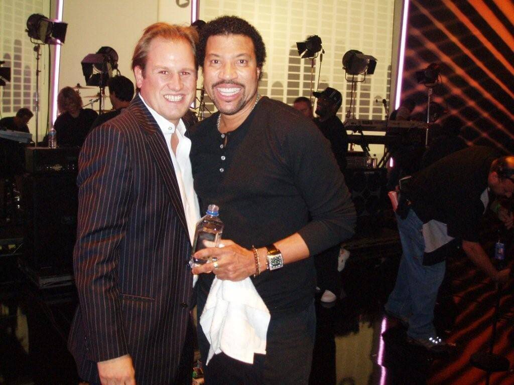 LiveTube - Two men stand together smiling at the camera in a studio setting; one holds a water bottle and a white towel. Studio lights and crew are visible in the background.
