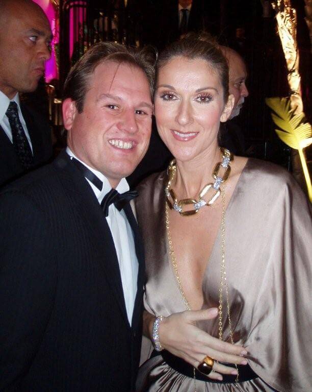 LiveTube - A man in a tuxedo poses with a woman in a brown dress and gold necklace at a formal event. Other people and decorative lighting are visible in the background.