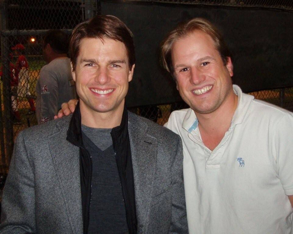 LiveTube - Two men are smiling and standing together in front of a chain-link fence, with one man’s arm around the other's shoulder.