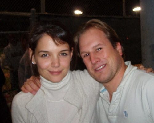 LiveTube - A woman and a man pose closely together and smile at the camera in an outdoor nighttime setting with a chain-link fence in the background.