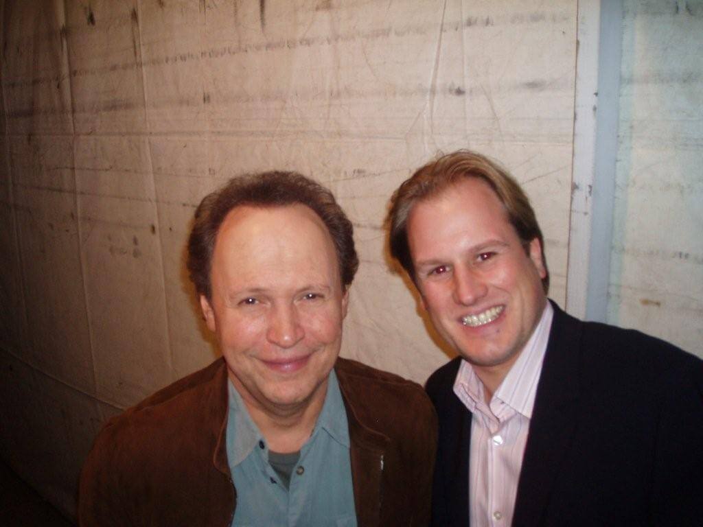 LiveTube - Two men standing together indoors, smiling at the camera. The background is a plain, light-colored wall.