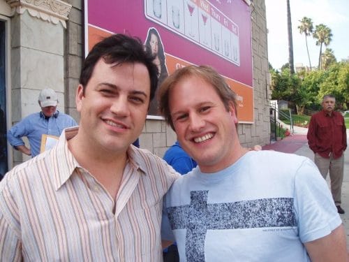 LiveTube - Two men are standing close together and smiling at the camera outside, with a billboard and other people visible in the background.