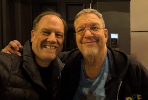 LiveTube - Two smiling men posing closely together for a photo, one with glasses and a blue shirt, the other wearing a black jacket, standing in front of a dark doorway.