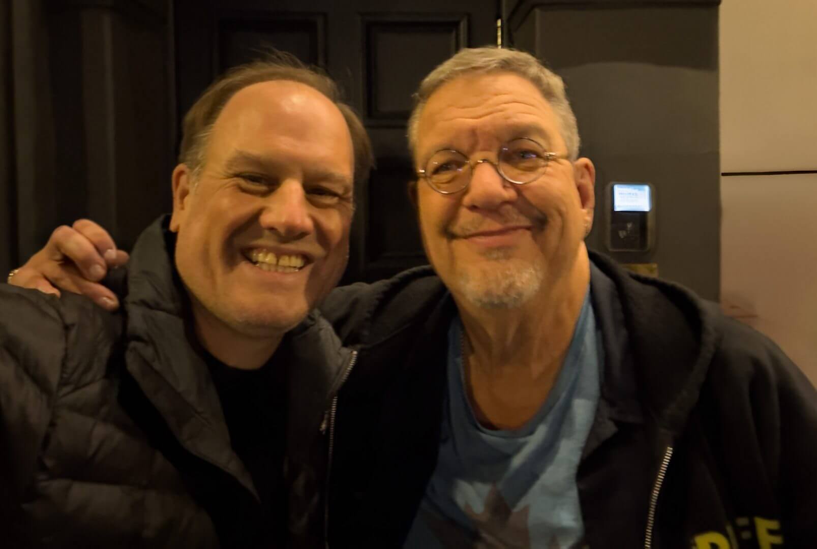 LiveTube - Two smiling men posing closely together for a photo, one with glasses and a blue shirt, the other wearing a black jacket, standing in front of a dark doorway.