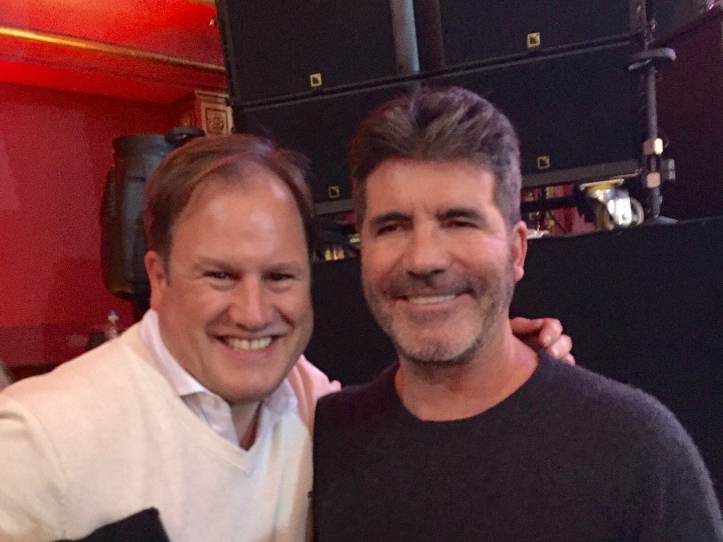 LiveTube - Two men are smiling and posing together indoors, with one wearing a white sweater and the other in a dark top. The background is dimly lit with equipment and red walls visible.