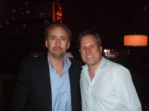 LiveTube - Two men wearing button-up shirts pose together and smile for the camera in a dimly lit indoor setting.