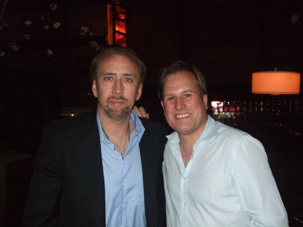 LiveTube - Two men wearing button-up shirts pose together and smile for the camera in a dimly lit indoor setting.