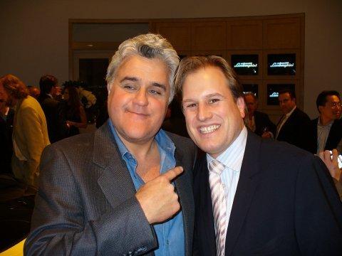 LiveTube - Two men in suits smile for a photo at an indoor event; one man is pointing at the other. People and screens are visible in the background.