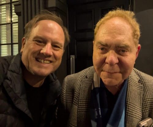 LiveTube - Two men standing close together and smiling at the camera, one wearing a quilted jacket and the other in a plaid blazer and scarf, in front of a dark door and window.